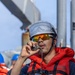 A maritime or naval worker is speaking into a corded radio handset while wearing full safety gear including a white hard hat, orange reflective life vest, and mirrored orange-tinted safety glasses. The setting appears to be the deck of a large vessel or port facility, with other personnel visible in the background also wearing safety equipment. The image captures an active communication moment during what appears to be a maritime operation or exercise.