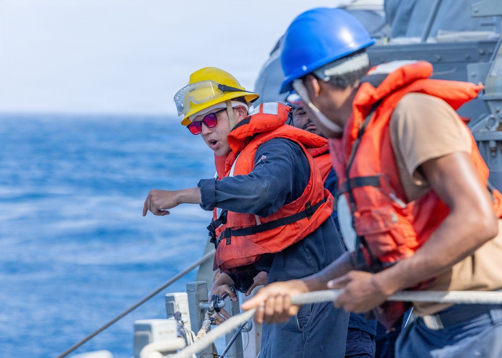 Three personnel are visible on the deck of a naval vessel at sea, wearing orange life vests and hard hats of different colors (yellow and blue). The individual in the yellow helmet appears to be giving directions or instructions while pointing, suggesting a coordinated deck operation. A rope or line is visible along the ship's railing, indicating a mooring, towing, or replenishment-at-sea activity.
