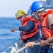 Three personnel are visible on the deck of a naval vessel at sea, wearing orange life vests and hard hats of different colors (yellow and blue). The individual in the yellow helmet appears to be giving directions or instructions while pointing, suggesting a coordinated deck operation. A rope or line is visible along the ship's railing, indicating a mooring, towing, or replenishment-at-sea activity.