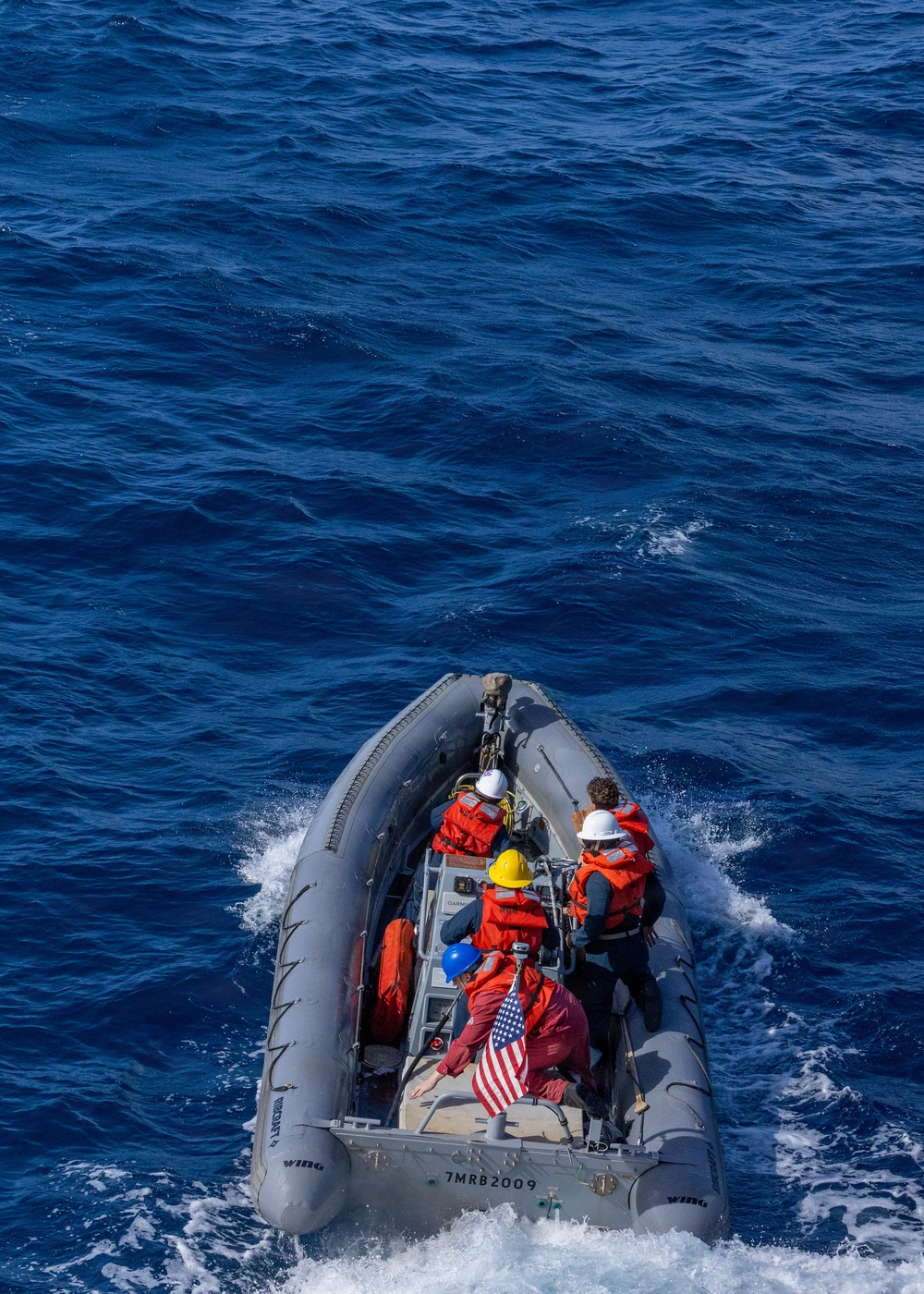 An aerial view of a rigid inflatable boat (RIB) designated '7MRB2009' underway on open blue ocean water. Four personnel are aboard, wearing orange immersion suits and colored hard hats, with an American flag visible on the vessel. The boat appears to be a military or coast guard craft based on its gray color scheme and markings.