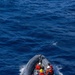 An aerial view of a rigid inflatable boat (RIB) designated '7MRB2009' underway on open blue ocean water. Four personnel are aboard, wearing orange immersion suits and colored hard hats, with an American flag visible on the vessel. The boat appears to be a military or coast guard craft based on its gray color scheme and markings.