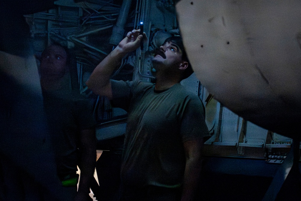 A person wearing a military-style olive green t-shirt is using a small penlight or tactical flashlight to inspect the underside of what appears to be a military aircraft in a dimly lit environment. The blue LED light illuminates the person's face from above as they look upward. At least one other individual is partially visible in the background.