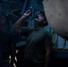 A person wearing a military-style olive green t-shirt is using a small penlight or tactical flashlight to inspect the underside of what appears to be a military aircraft in a dimly lit environment. The blue LED light illuminates the person's face from above as they look upward. At least one other individual is partially visible in the background.