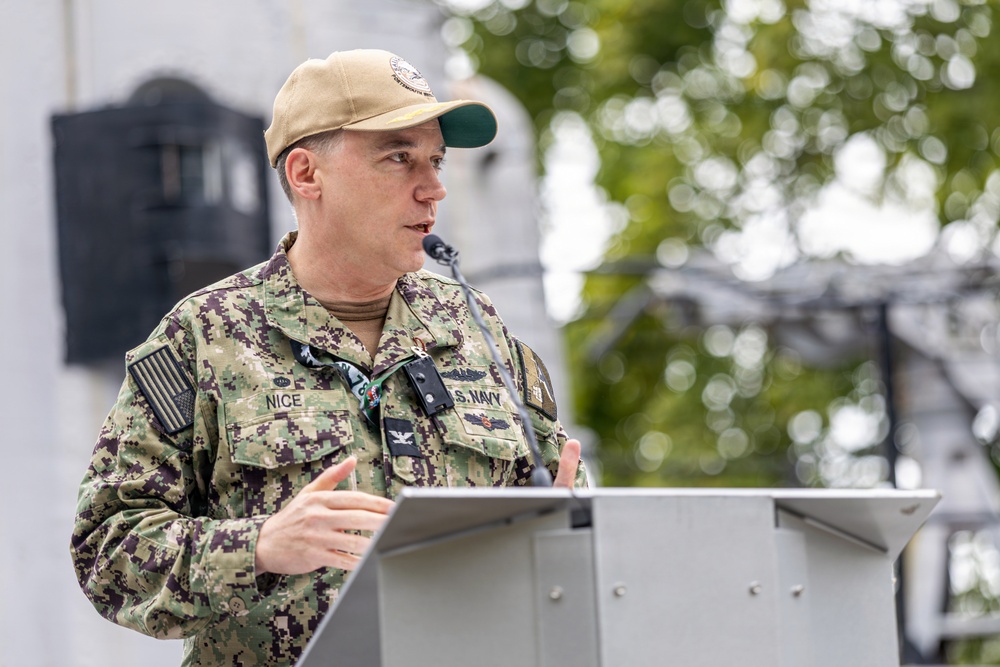 A U.S. Navy officer in NWU Type III (digital camouflage) uniform is speaking at a podium outdoors. The name tape reads 'NICE' and the branch tape reads 'U.S. NAVY', with visible rank insignia of a Captain (O-6, eagle device) on the collar. The setting appears to be an official military ceremony or briefing.