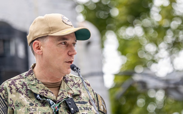A U.S. Navy officer in NWU Type III (digital camouflage) uniform is speaking at a podium outdoors. The name tape reads 'NICE' and the branch tape reads 'U.S. NAVY', with visible rank insignia of a Captain (O-6, eagle device) on the collar. The setting appears to be an official military ceremony or briefing.