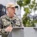 A U.S. Navy officer in NWU Type III (digital camouflage) uniform is speaking at a podium outdoors. The name tape reads 'NICE' and the branch tape reads 'U.S. NAVY', with visible rank insignia of a Captain (O-6, eagle device) on the collar. The setting appears to be an official military ceremony or briefing.