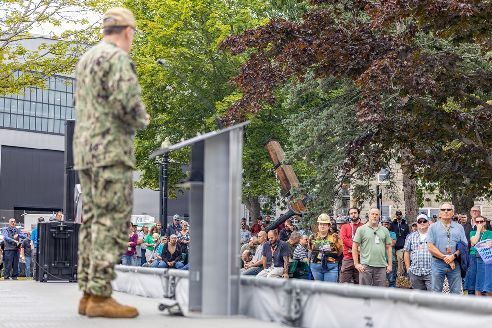 A U.S. Navy officer in camouflage uniform stands at a podium addressing a large crowd gathered outdoors. The event appears to be a formal military ceremony or public address, taking place in an open plaza area surrounded by trees and buildings. A diverse crowd of civilians and workers, some wearing hard hats and lanyards, attentively listens to the speaker.