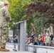 A U.S. Navy officer in camouflage uniform stands at a podium addressing a large crowd gathered outdoors. The event appears to be a formal military ceremony or public address, taking place in an open plaza area surrounded by trees and buildings. A diverse crowd of civilians and workers, some wearing hard hats and lanyards, attentively listens to the speaker.