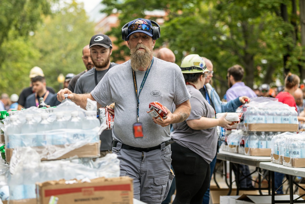 A group of workers or volunteers are distributing supplies, primarily cases of Polar brand bottled water, at what appears to be an outdoor relief or supply distribution event. The central figure is a bearded man wearing a blue cap, headphones, and a lanyard with an ID badge, carrying a pack of water bottles and a small box. The scene suggests an organized community or emergency response effort taking place in an urban outdoor setting.