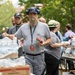 A group of workers or volunteers are distributing supplies, primarily cases of Polar brand bottled water, at what appears to be an outdoor relief or supply distribution event. The central figure is a bearded man wearing a blue cap, headphones, and a lanyard with an ID badge, carrying a pack of water bottles and a small box. The scene suggests an organized community or emergency response effort taking place in an urban outdoor setting.