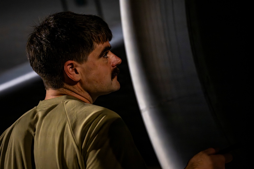 A man with dark hair and a mustache is photographed in profile, wearing an olive/military green shirt. He appears to be inspecting or working on a large curved metallic or composite surface, likely an aircraft component. The image is dramatically lit with strong contrast between light and shadow.