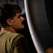A man with dark hair and a mustache is photographed in profile, wearing an olive/military green shirt. He appears to be inspecting or working on a large curved metallic or composite surface, likely an aircraft component. The image is dramatically lit with strong contrast between light and shadow.