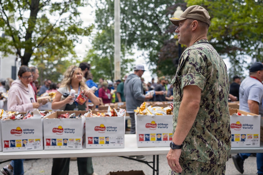 An outdoor food distribution event is taking place, with a U.S. Navy service member in digital camouflage (NWU Type III) standing near tables stocked with Frito-Lay 60 Bags Variety snack boxes. Civilians and volunteers are actively distributing snack bags to a crowd of people gathered around the tables. The setting appears to be a public outdoor area with trees visible in the background.