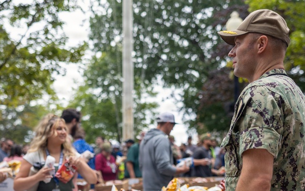 An outdoor food distribution event is taking place, with a U.S. Navy service member in digital camouflage (NWU Type III) standing near tables stocked with Frito-Lay 60 Bags Variety snack boxes. Civilians and volunteers are actively distributing snack bags to a crowd of people gathered around the tables. The setting appears to be a public outdoor area with trees visible in the background.