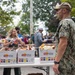 An outdoor food distribution event is taking place, with a U.S. Navy service member in digital camouflage (NWU Type III) standing near tables stocked with Frito-Lay 60 Bags Variety snack boxes. Civilians and volunteers are actively distributing snack bags to a crowd of people gathered around the tables. The setting appears to be a public outdoor area with trees visible in the background.