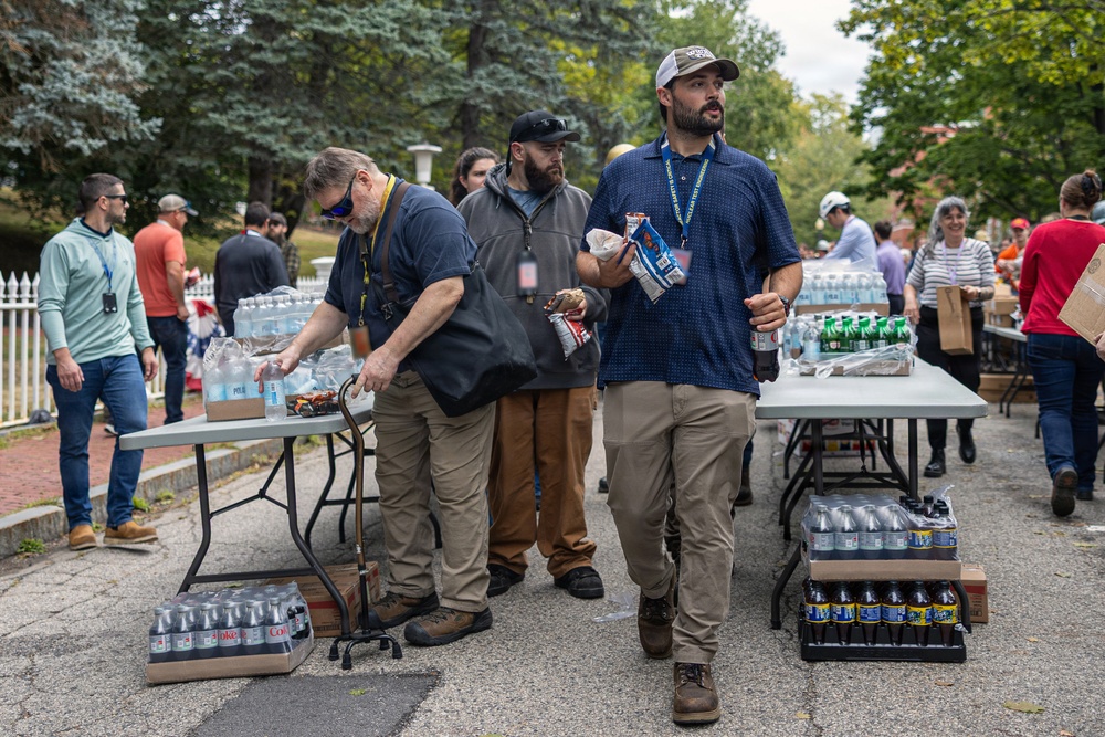 An outdoor distribution or relief event is taking place, with multiple people gathered around folding tables stocked with bottled water, sodas, and snacks. Several individuals wearing lanyards appear to be volunteers or organizers handing out supplies. The setting appears to be a paved area near a tree-lined street or park.