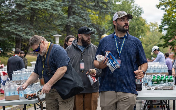 An outdoor distribution or relief event is taking place, with multiple people gathered around folding tables stocked with bottled water, sodas, and snacks. Several individuals wearing lanyards appear to be volunteers or organizers handing out supplies. The setting appears to be a paved area near a tree-lined street or park.