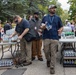 An outdoor distribution or relief event is taking place, with multiple people gathered around folding tables stocked with bottled water, sodas, and snacks. Several individuals wearing lanyards appear to be volunteers or organizers handing out supplies. The setting appears to be a paved area near a tree-lined street or park.