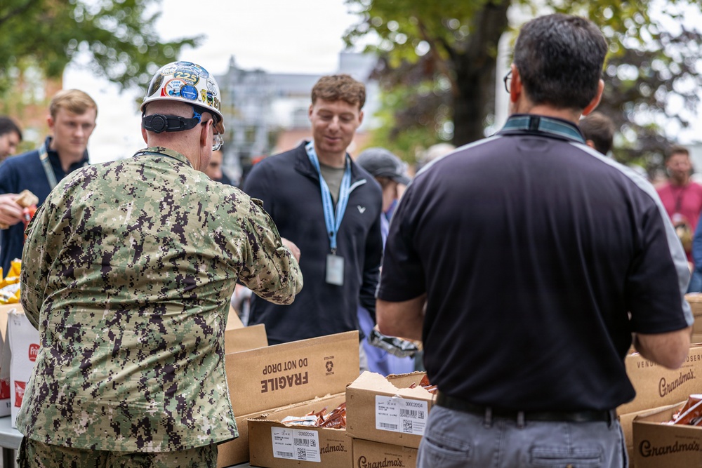 A group of people are gathered outdoors around tables stacked with cardboard boxes, appearing to be involved in a distribution or packing event. One individual is wearing a U.S. Navy digital camouflage uniform and a hard hat decorated with stickers, while others are in civilian clothing. The boxes visible include 'Grandma's' branded packaging and boxes marked 'FRAGILE - DO NOT DROP.'