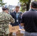A group of people are gathered outdoors around tables stacked with cardboard boxes, appearing to be involved in a distribution or packing event. One individual is wearing a U.S. Navy digital camouflage uniform and a hard hat decorated with stickers, while others are in civilian clothing. The boxes visible include 'Grandma's' branded packaging and boxes marked 'FRAGILE - DO NOT DROP.'