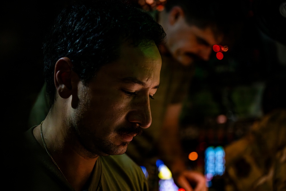 A close-up, low-light photograph of a young man with short dark hair and facial stubble, looking downward in a contemplative pose. He is wearing a green t-shirt and a thin chain necklace. A second person is visible in the blurred background, along with colorful bokeh lights suggesting a nighttime or indoor entertainment environment.