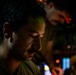 A close-up, low-light photograph of a young man with short dark hair and facial stubble, looking downward in a contemplative pose. He is wearing a green t-shirt and a thin chain necklace. A second person is visible in the blurred background, along with colorful bokeh lights suggesting a nighttime or indoor entertainment environment.