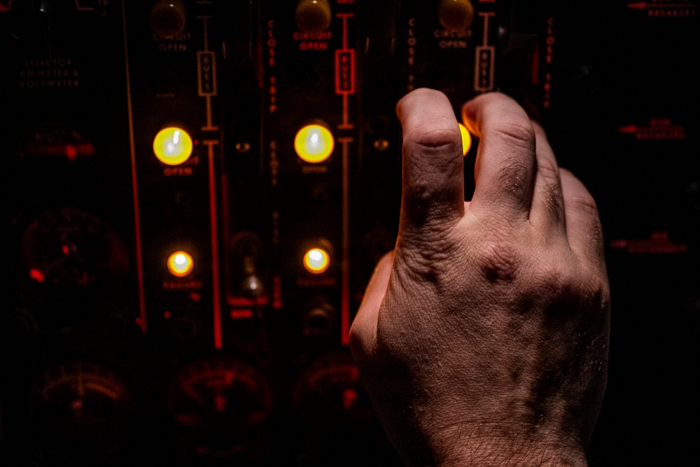 A human hand is reaching toward or operating a dark industrial control panel featuring illuminated yellow indicator lights and labeled switches. The panel displays text including 'CIRCUIT OPEN' and 'CLOSE' markings, suggesting electrical switching equipment. The image is dramatically lit with warm amber tones from the glowing indicators against a dark background.
