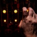 A human hand is reaching toward or operating a dark industrial control panel featuring illuminated yellow indicator lights and labeled switches. The panel displays text including 'CIRCUIT OPEN' and 'CLOSE' markings, suggesting electrical switching equipment. The image is dramatically lit with warm amber tones from the glowing indicators against a dark background.