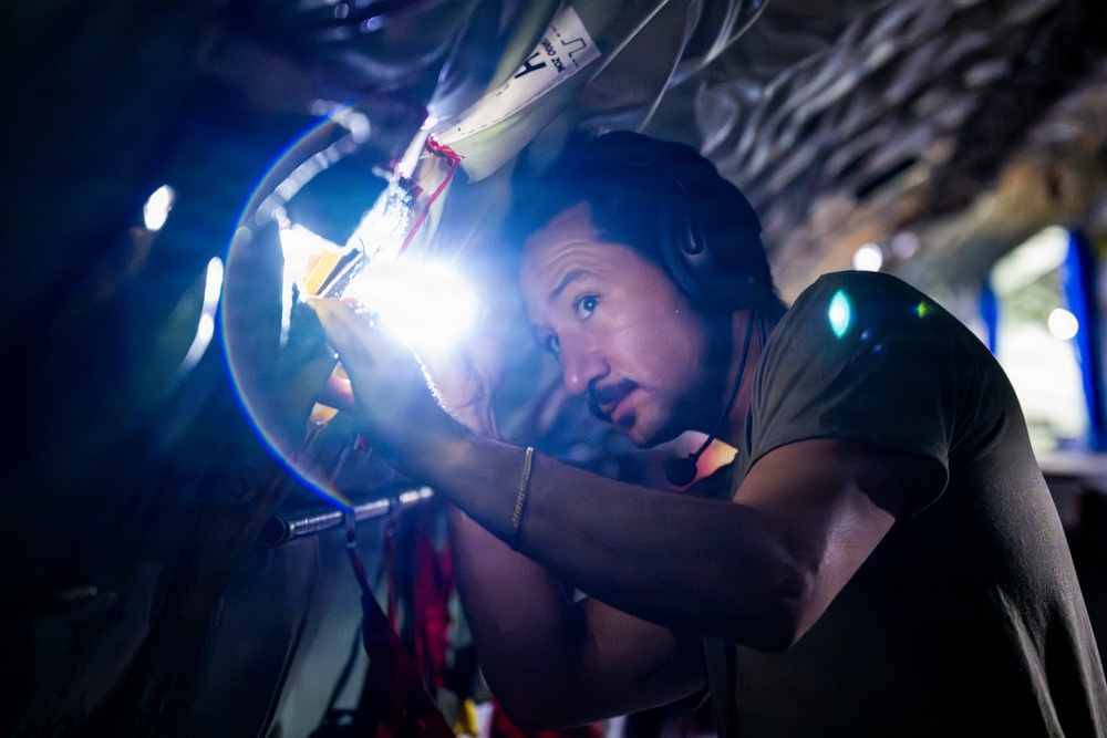 A man wearing headphones and a dark t-shirt is closely inspecting or working on a mechanical or structural component using a bright flashlight or work light. He appears to be in a confined, dimly lit space, possibly inside a vehicle, aircraft, or industrial structure. The bright light source creates strong contrast and lens flare in the image.
