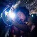 A man wearing headphones and a dark t-shirt is closely inspecting or working on a mechanical or structural component using a bright flashlight or work light. He appears to be in a confined, dimly lit space, possibly inside a vehicle, aircraft, or industrial structure. The bright light source creates strong contrast and lens flare in the image.