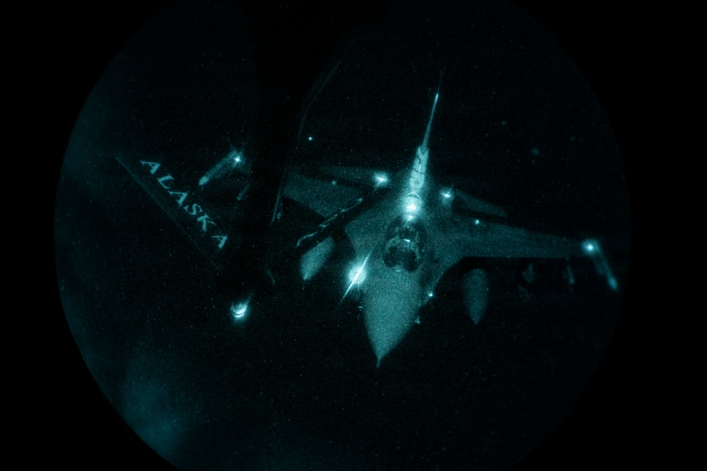 A night-vision or infrared image captured through a circular optical device (likely a boom operator's window or night-vision optic) showing a military aircraft during an aerial refueling operation. The word 'ALASKA' is visible on what appears to be a tanker aircraft's fuselage or wing. Multiple navigation and position lights are illuminated on the aircraft(s) visible in the frame.