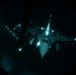 A night-vision or infrared image captured through a circular optical device (likely a boom operator's window or night-vision optic) showing a military aircraft during an aerial refueling operation. The word 'ALASKA' is visible on what appears to be a tanker aircraft's fuselage or wing. Multiple navigation and position lights are illuminated on the aircraft(s) visible in the frame.