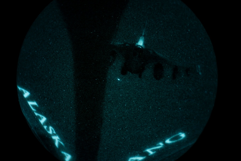 A night-vision or thermal imaging view through what appears to be a circular optical device (likely a night-vision goggle or scope) showing a military aircraft in flight. The image has a characteristic teal/green monochromatic appearance typical of night-vision equipment. Text reading 'ALASKA' and partial numbers/letters are visible along the curved edge of the viewing aperture.