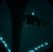 A night-vision or thermal imaging view through what appears to be a circular optical device (likely a night-vision goggle or scope) showing a military aircraft in flight. The image has a characteristic teal/green monochromatic appearance typical of night-vision equipment. Text reading 'ALASKA' and partial numbers/letters are visible along the curved edge of the viewing aperture.