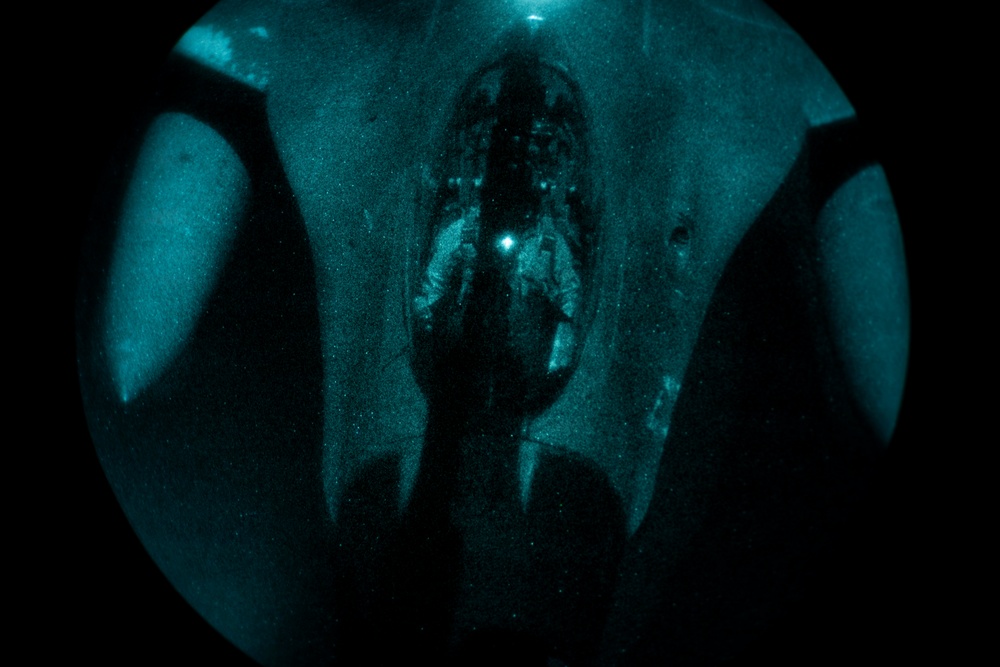 The image appears to be captured through a night vision or image intensifier device, producing a characteristic circular green-teal tinted view. The scene shows what appears to be the interior of a large military aircraft, likely viewed through the open rear cargo ramp or door, with at least one figure visible inside. The low-light imaging technology creates a grainy, monochromatic teal appearance typical of night vision optics.