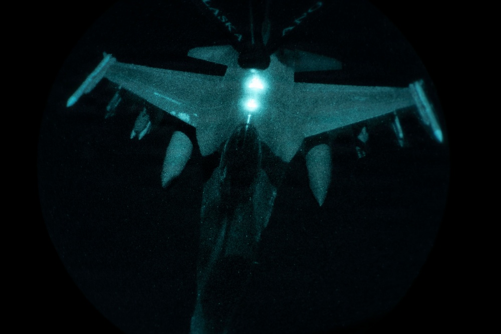 A night aerial refueling operation captured from the boom operator's perspective aboard a tanker aircraft. A fighter jet, consistent with an F-16 Fighting Falcon configuration, is visible below receiving or approaching the refueling boom. The image is illuminated by the tanker's refueling lights, casting a teal/blue-green glow against an otherwise completely dark background.