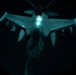 A night aerial refueling operation captured from the boom operator's perspective aboard a tanker aircraft. A fighter jet, consistent with an F-16 Fighting Falcon configuration, is visible below receiving or approaching the refueling boom. The image is illuminated by the tanker's refueling lights, casting a teal/blue-green glow against an otherwise completely dark background.