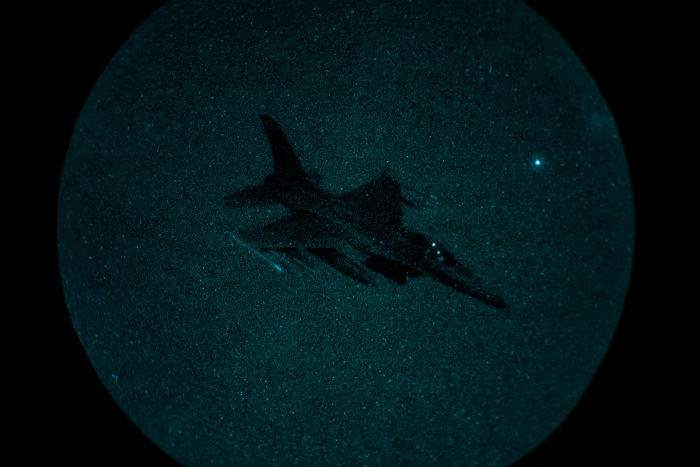 A night vision image captured through a circular optical device (likely night vision goggles or a scope) showing a military jet aircraft in flight. The image displays the characteristic green-teal phosphor glow typical of image intensifier night vision technology. The aircraft silhouette is clearly visible against a grainy, star-speckled night sky.