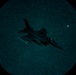 A night vision image captured through a circular optical device (likely night vision goggles or a scope) showing a military jet aircraft in flight. The image displays the characteristic green-teal phosphor glow typical of image intensifier night vision technology. The aircraft silhouette is clearly visible against a grainy, star-speckled night sky.