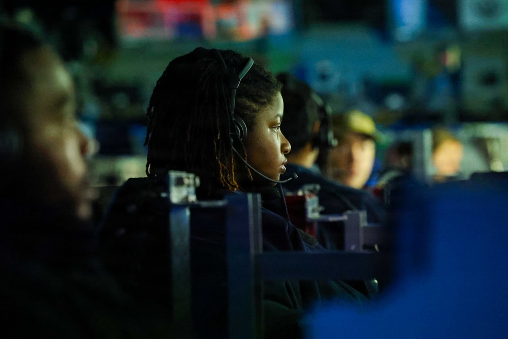 The image shows a focused individual with dreadlocks wearing a headset, seated in what appears to be a dimly lit gaming or esports event environment. Multiple participants are visible in the background, similarly equipped with headsets and seated at stations. The shallow depth of field keeps the central subject sharp while the background remains blurred.