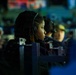 The image shows a focused individual with dreadlocks wearing a headset, seated in what appears to be a dimly lit gaming or esports event environment. Multiple participants are visible in the background, similarly equipped with headsets and seated at stations. The shallow depth of field keeps the central subject sharp while the background remains blurred.
