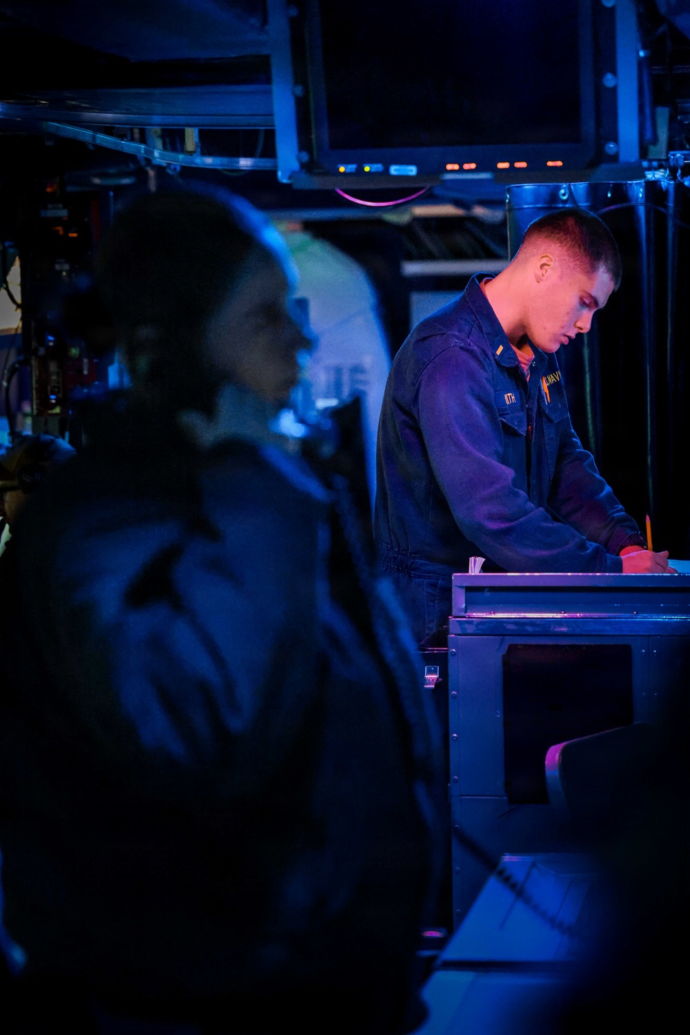 Two military personnel are visible in what appears to be a ship's combat information center or bridge, illuminated by blue ambient lighting. The sailor in focus is wearing a dark navy coverall uniform and is leaning over a console or workstation, appearing to review documents or operate equipment. Multiple overhead display screens are visible in the background, consistent with a naval vessel's operational spaces.