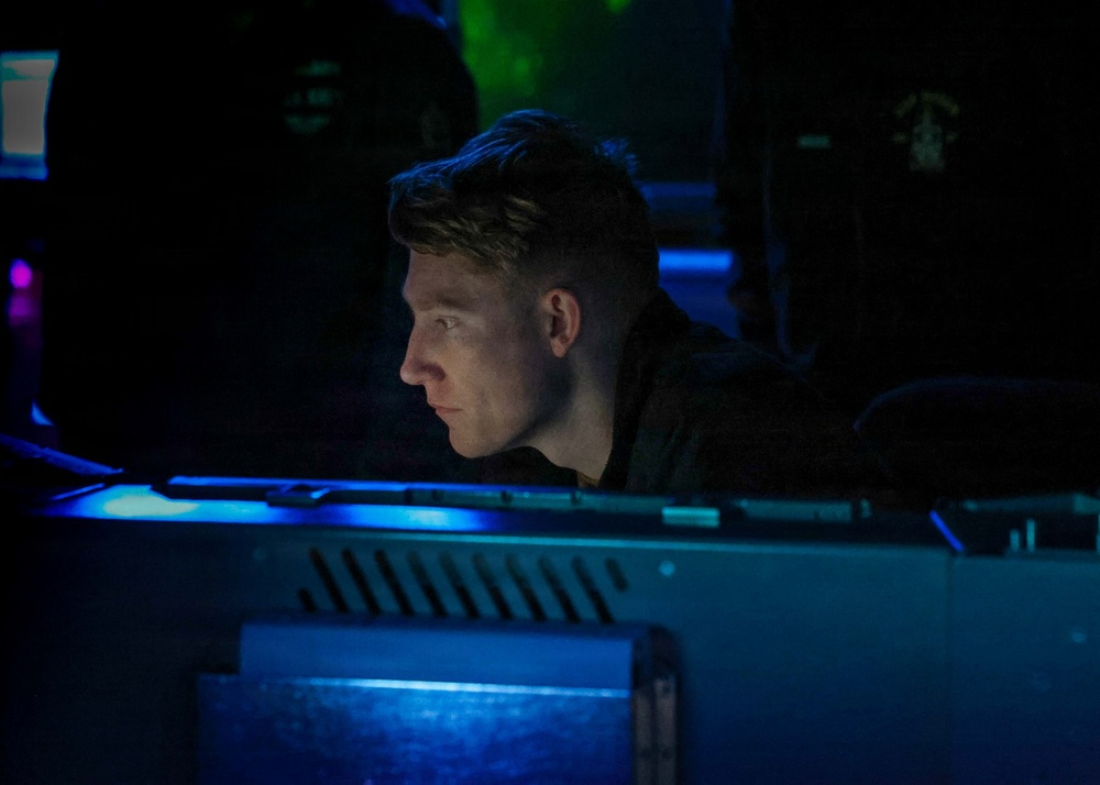 A young man is seated at what appears to be a darkened operations or control room, illuminated primarily by blue light emanating from monitors or equipment in front of him. His face is lit from below by the screen glow as he focuses intently on the display. The environment suggests a military, naval, or technical operations setting.