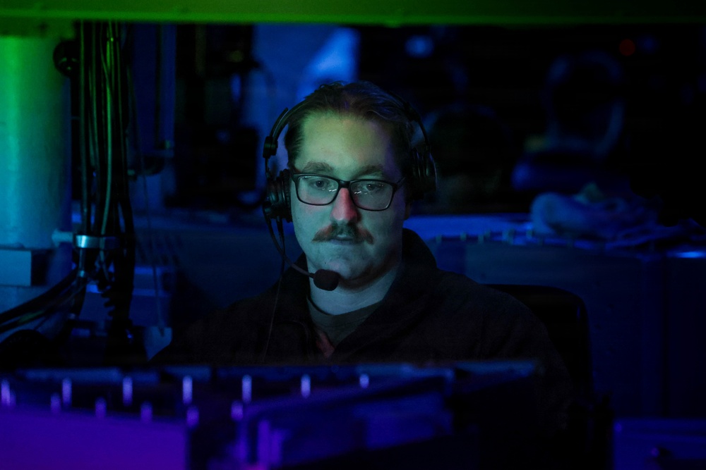 A man wearing glasses and a headset with a boom microphone is seated at what appears to be a workstation in a darkened room illuminated by blue and green ambient lighting. He is focused on something in front of him, likely a monitor or control panel. The setting suggests a gaming, broadcasting, or technical operations environment.