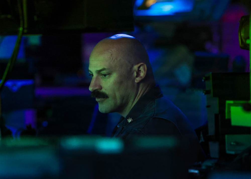 A bald man with a mustache is seated at what appears to be a dimly lit operations or command center, illuminated by blue and green ambient lighting from surrounding screens and equipment. He is wearing a dark uniform with a small pin or insignia visible on his chest. The environment suggests a military, naval, or technical operations setting.