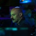 A bald man with a mustache is seated at what appears to be a dimly lit operations or command center, illuminated by blue and green ambient lighting from surrounding screens and equipment. He is wearing a dark uniform with a small pin or insignia visible on his chest. The environment suggests a military, naval, or technical operations setting.