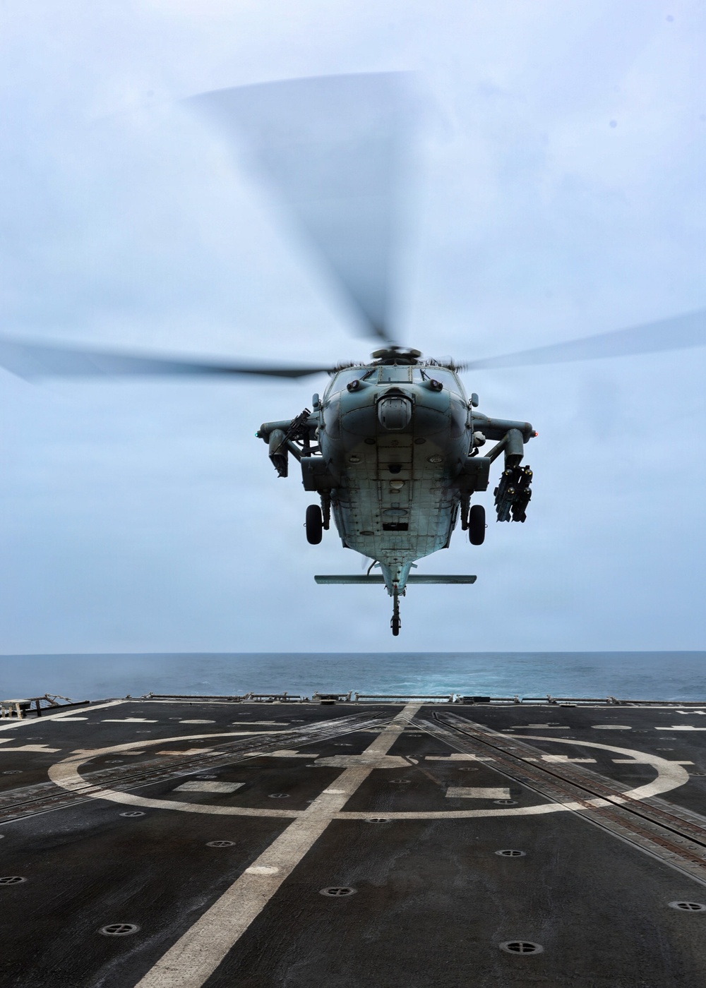 A military helicopter is hovering just above a naval vessel's flight deck over open ocean. The helicopter appears to be in the process of landing or taking off from the ship's helipad, which is marked with a circular landing zone and cross markings. The overcast sky and calm sea are visible in the background.