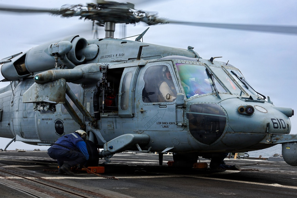 A U.S. Navy helicopter, numbered 610, is on the flight deck of what appears to be the USS George H.W. Bush aircraft carrier. The rotors are spinning, indicating the aircraft is either preparing for takeoff or has just landed. Ground crew personnel are working around the aircraft while pilots are visible in the cockpit.
