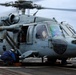 A U.S. Navy helicopter, numbered 610, is on the flight deck of what appears to be the USS George H.W. Bush aircraft carrier. The rotors are spinning, indicating the aircraft is either preparing for takeoff or has just landed. Ground crew personnel are working around the aircraft while pilots are visible in the cockpit.