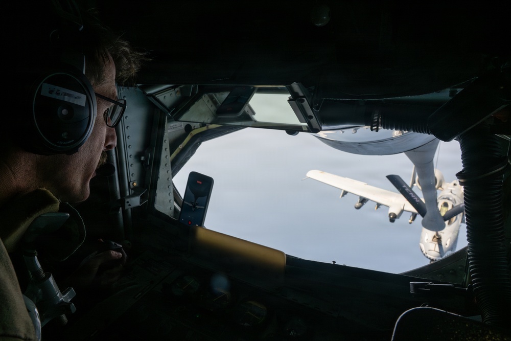 A boom operator aboard a tanker aircraft is observed monitoring an aerial refueling operation from the rear boom operator station. Through the observation window, a large multi-engine aircraft (appearing to be a C-130 or similar transport) is visible receiving or approaching for fuel transfer via a refueling boom. The interior is dimly lit, contrasting with the bright sky visible through the window.